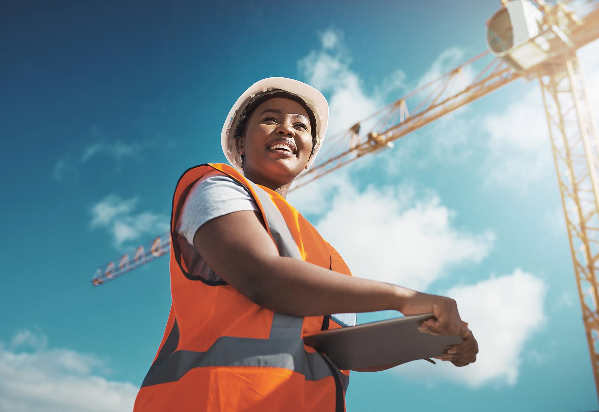 A smiling construction worker in a hard hat and safety vest holding a tablet, with a crane and blue sky behind her